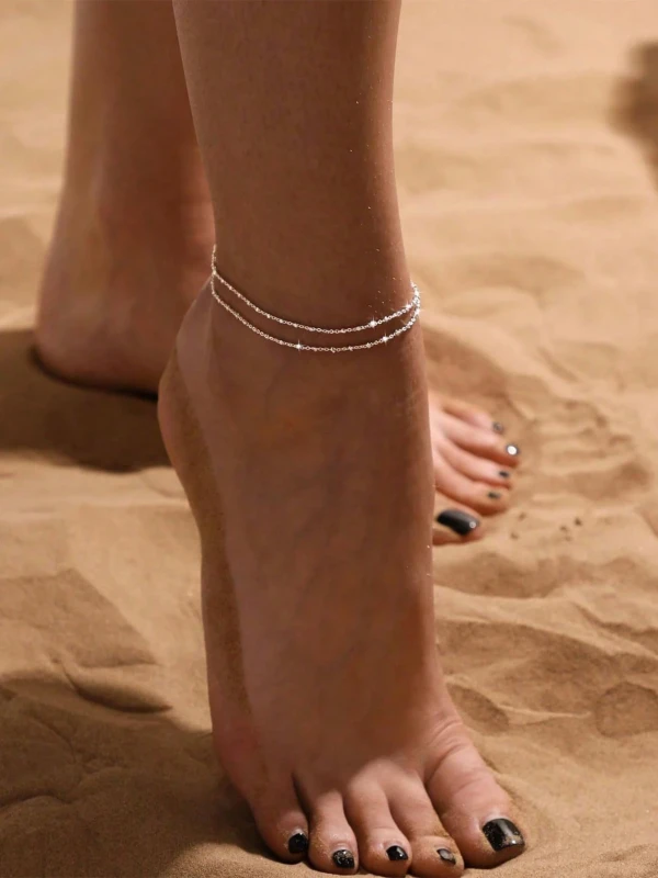 Srebrna podwójna bransoletka na nogę z drobnym połyskiem, noszona na kostce na plaży, przy sandałowym piasku (2).webp