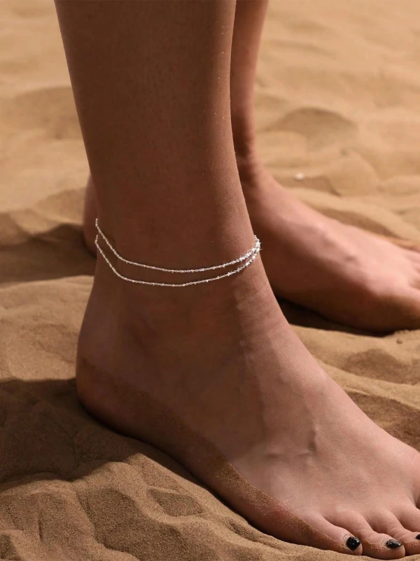 Srebrna podwójna bransoletka na nogę z drobnym połyskiem, noszona na kostce na plaży, przy sandałowym piasku (1).webp