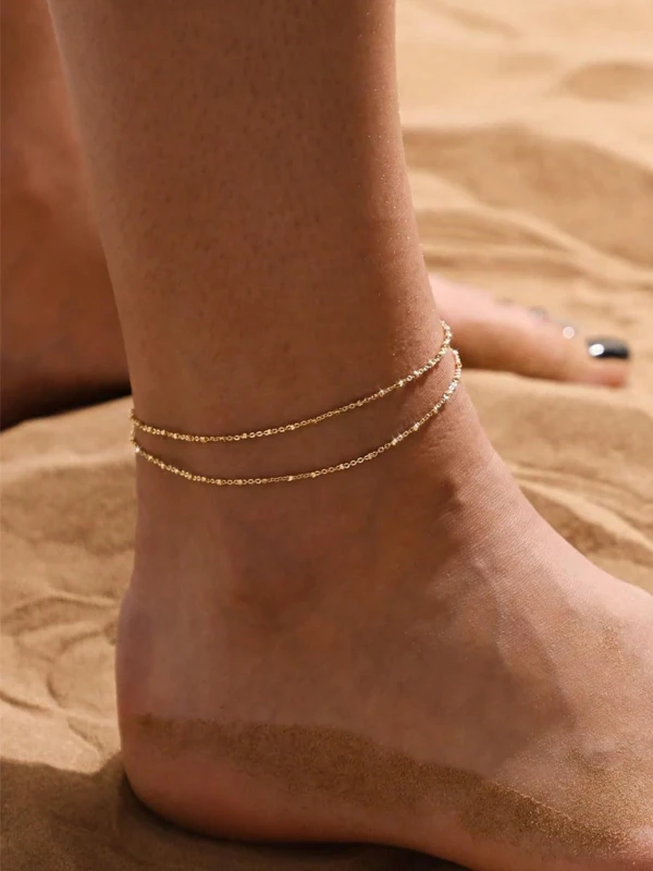 Złota podwójna bransoletka na nogę z drobnym połyskiem, noszona na kostce na plaży, na tle piasku (4).webp